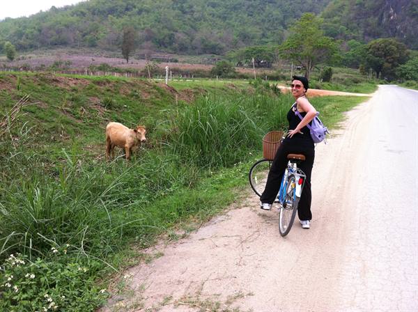 Woman with bicycle beside a calf grazing along a dirt road