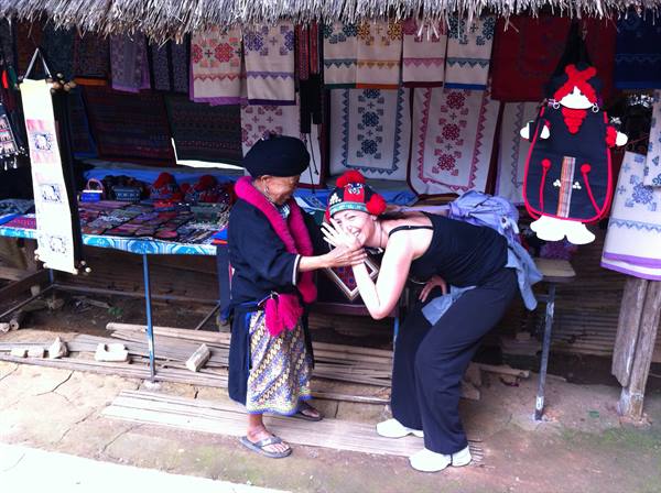 Akha woman in traditional dress caresses a tourist's face under a textile stall