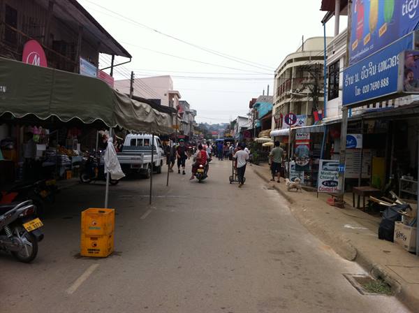 Busy market street with awnings, scooters and pedestrians under grey sky, Lao signs