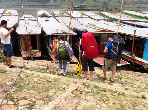 Backpackers walking down stone steps toward wooden riverboats moored at the shore