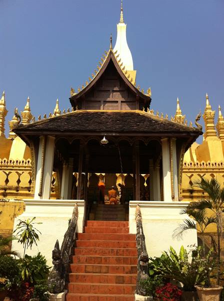 Orange-robed monk climbs stairs of golden Pha That Luang stupa under blue sky