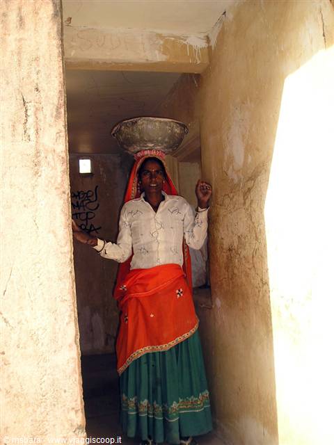 Woman in red and green sari balancing a metal basin on her head in a corridor