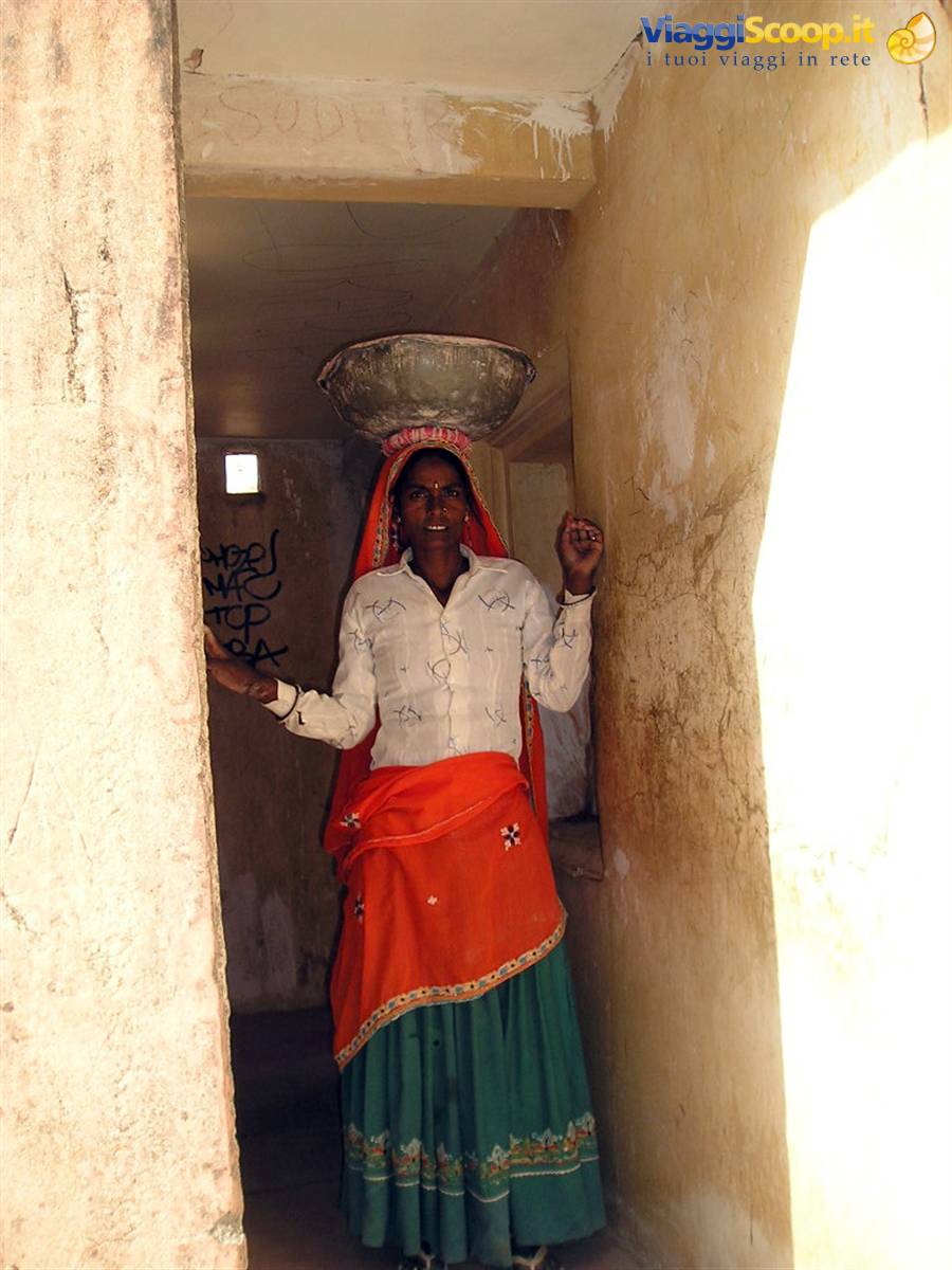 Woman in red and green sari balancing a metal basin on her head in a corridor