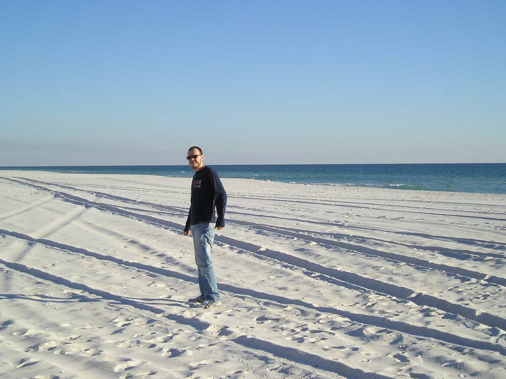 Man in sunglasses walking on white sand with tire tracks, turquoise sea behind