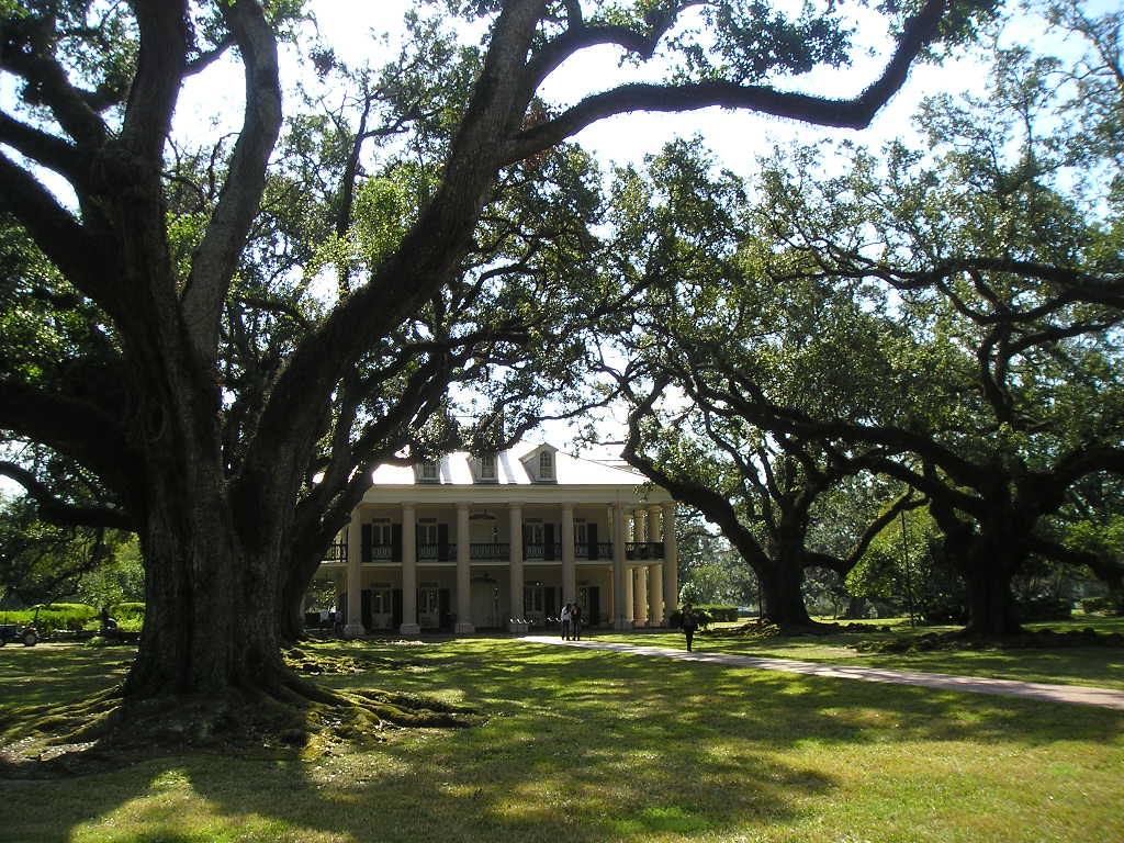 White columned plantation mansion shaded by giant oaks, visitors walk on the lawn