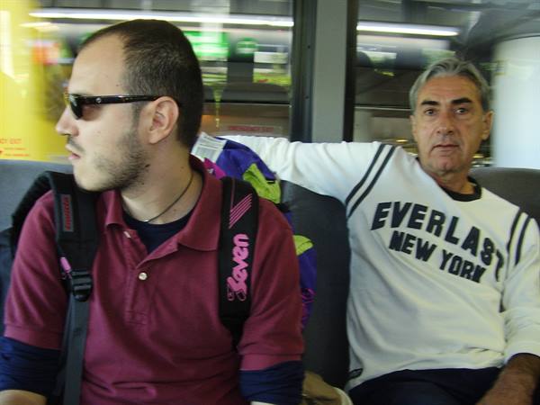Two men seated on a bus, one with backpack and sunglasses, one in Everlast sweatshirt