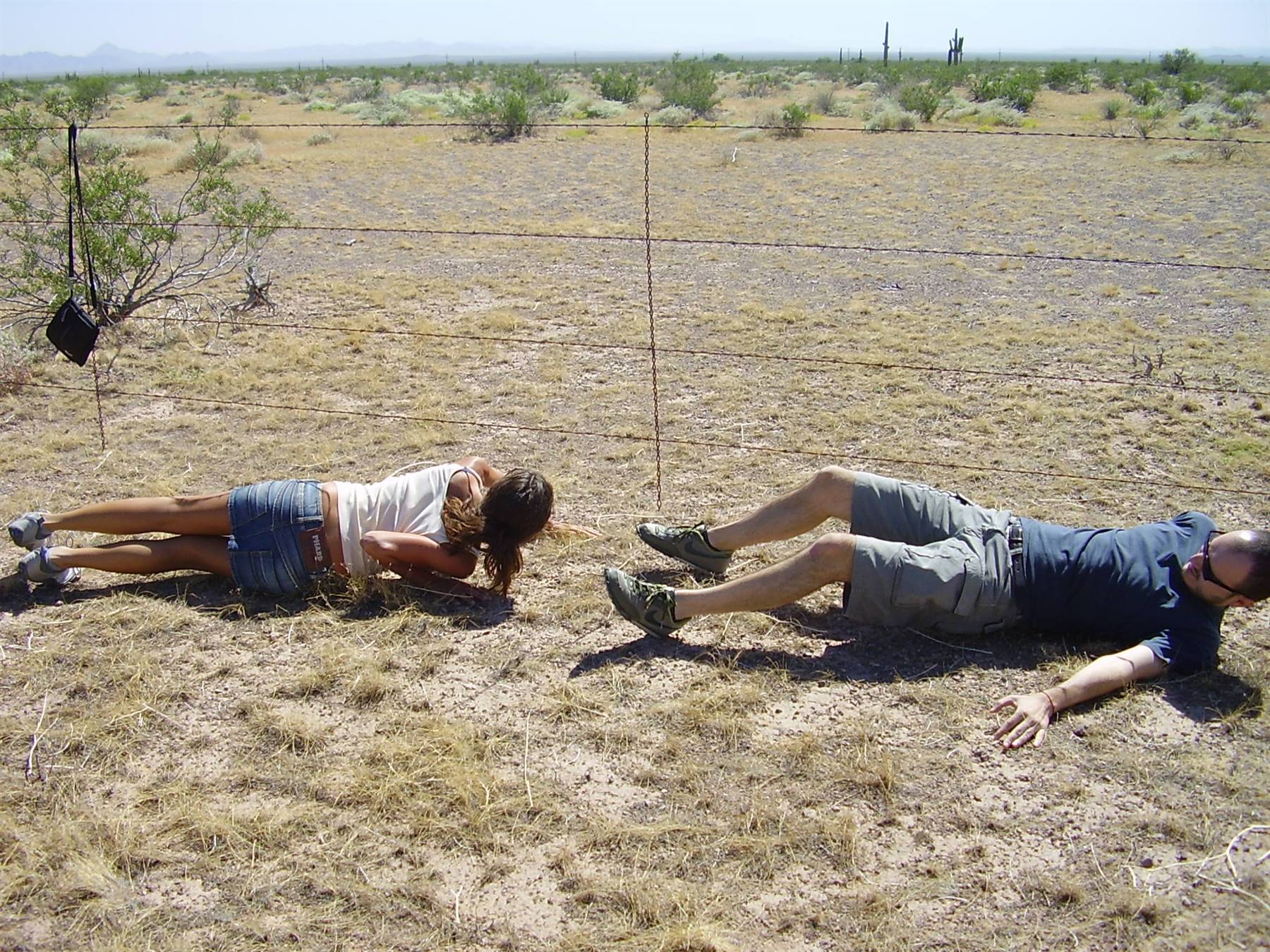 Two tourists exercising on dry desert ground, barbed wire fence and cacti behind