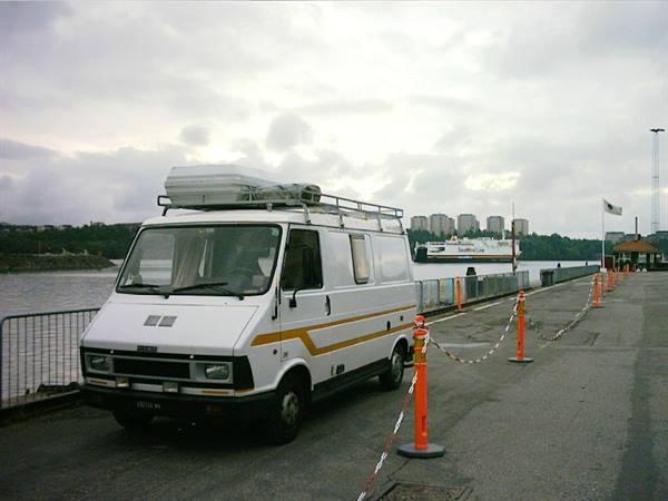 White camper van with roof luggage at a ferry dock, cloudy sky