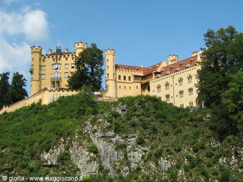 Yellow Hohenschwangau Castle with towers above rocky cliff and green trees