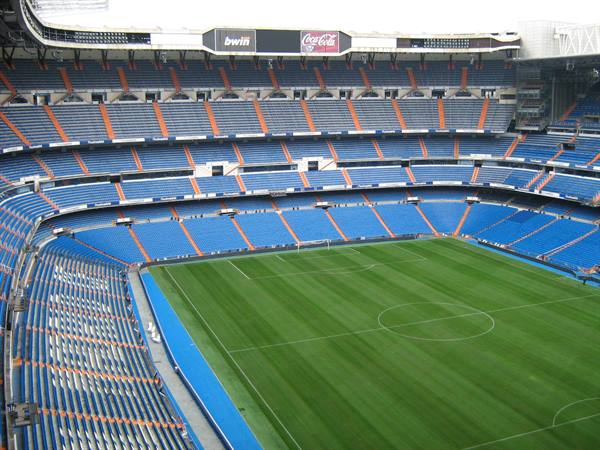 Empty Santiago Bernabéu stadium: green pitch, blue and orange stands under grey light