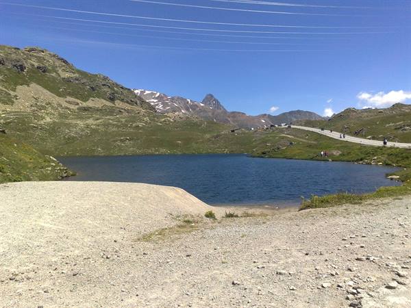 Alpine lake with gravel shore, cyclists on the pass road, snowy peaks
