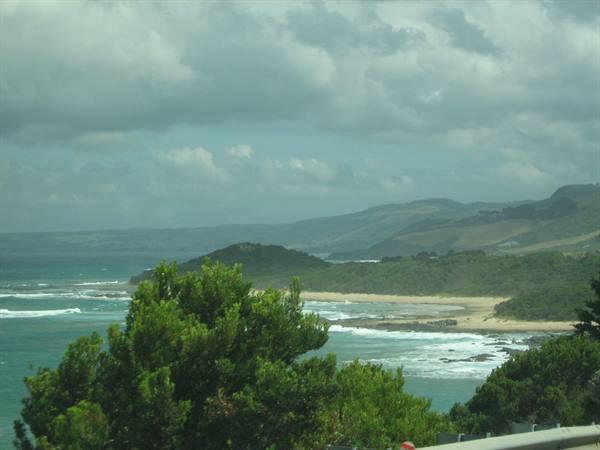Green trees before sandy beach and turquoise waves, hills under cloudy sky