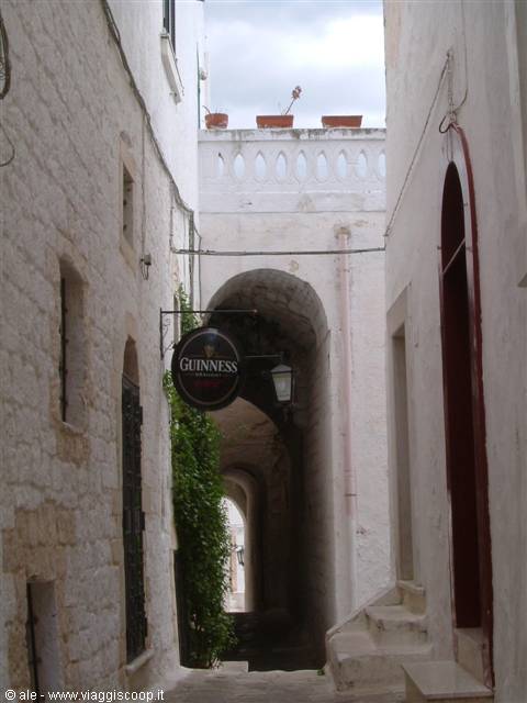 Alley with stone arches between whitewashed houses and a Guinness sign, Ostuni