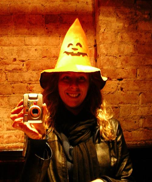 Woman in paper pumpkin hat smiles taking a photo before a brick wall