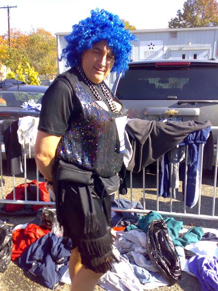 Man in blue wig and sequin vest by a barrier full of clothes