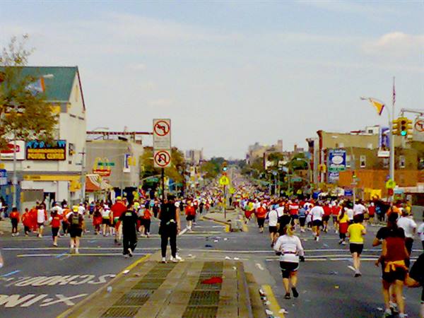 Runners fill a wide city street with crosswalks, colorful storefronts, hazy sky