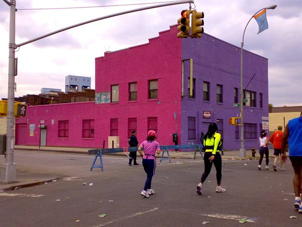 Runners cross the street by a pink and purple building, traffic light and blue barriers
