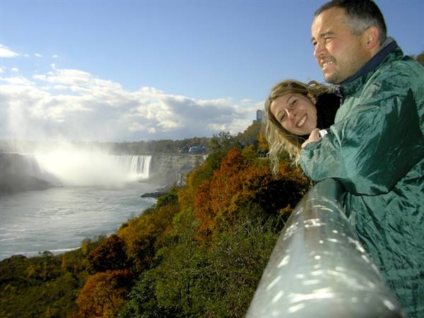 Couple at green railing before Niagara Falls, autumn foliage, sunny sky