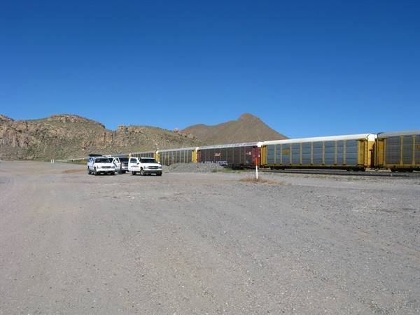 Treno merci giallo fermo sulla sterrata, montagne rocciose, 4x4 parcheggiati