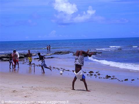Boy does a handstand on the sand while friends play football by wet rocks