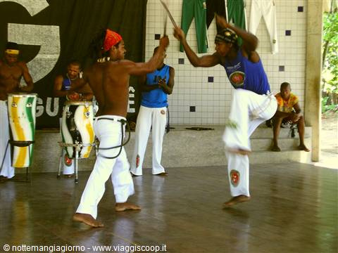 Two capoeiristas spar with sticks, atabaque drums behind them, Salvador de Bahia