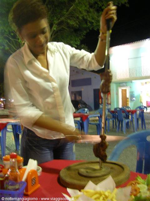 Waitress carves skewered meat onto wooden board at outdoor night eatery