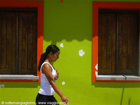 Woman walks past bright green wall with orange wooden window frames, Burano