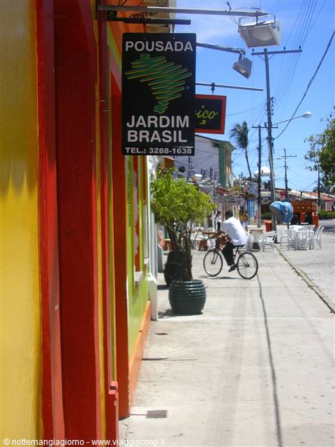 Pousada Jardim Brasil sign on yellow facade, a cyclist rides along sunny sidewalk