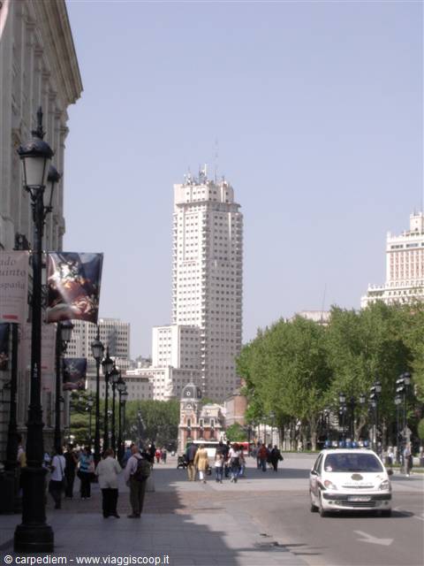 Pedestrians walking along a tree-lined street with the Torre de Madrid behind