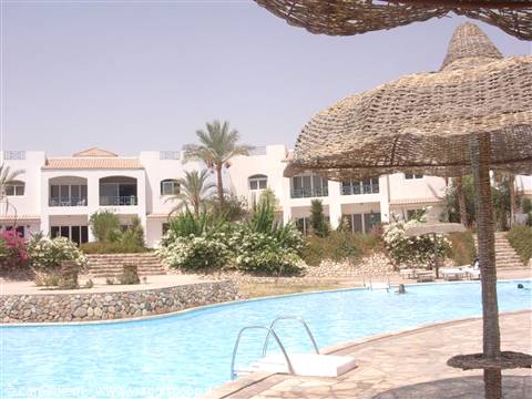 Blue pool with straw parasols and white hotel among palm trees