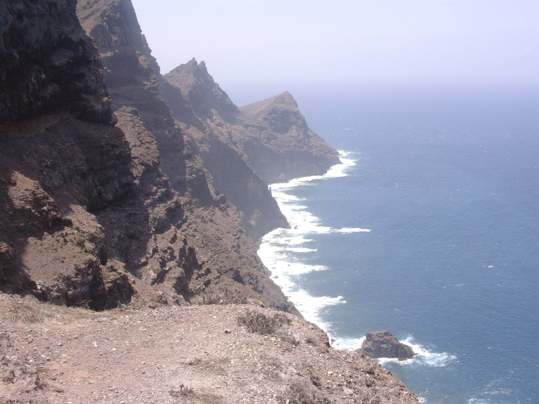 Brown rocky cliffs plunging into blue sea, white foam on waves