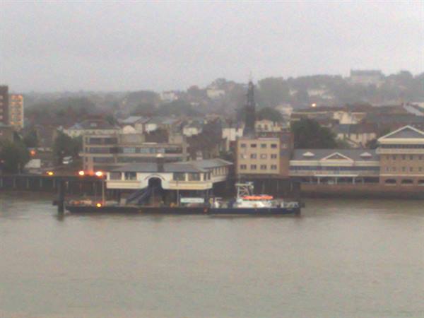 Ferry pier on grey river, rainy town buildings with church spire behind