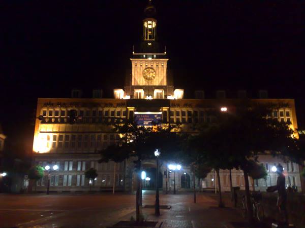 Illuminated town hall at night with clock tower and tree-lined square