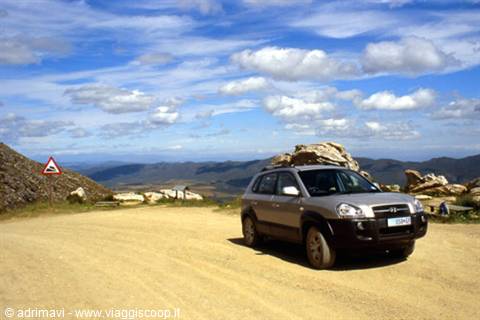 Grey SUV on mountain dirt road among rocks, cloudy sky