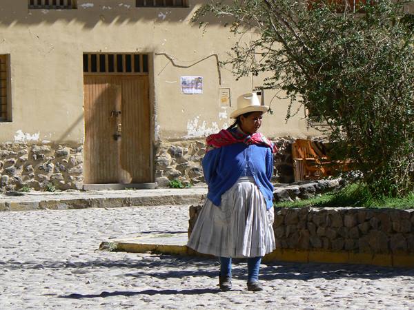 Woman in straw hat and gray skirt walks on cobblestone street by adobe house