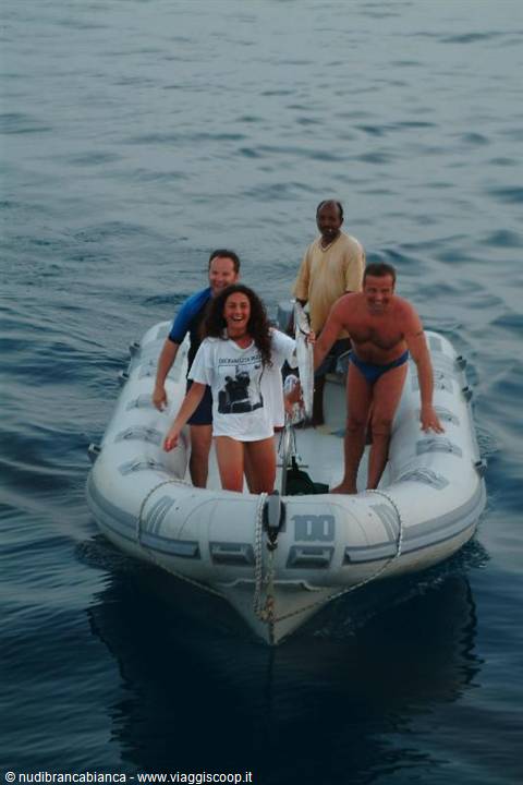 Four people on a grey inflatable dinghy on calm sea, smiling woman in white t-shirt