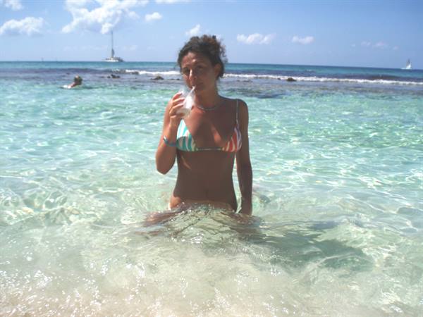 Woman in colorful bikini sips a drink standing in turquoise water, sailboats behind