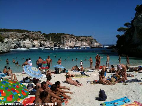 Beachgoers under colorful umbrellas on white sand, boats in Cala Macarella