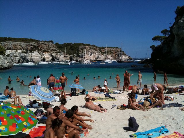 Bagnanti sotto ombrelloni colorati su sabbia bianca, barche in Cala Macarella