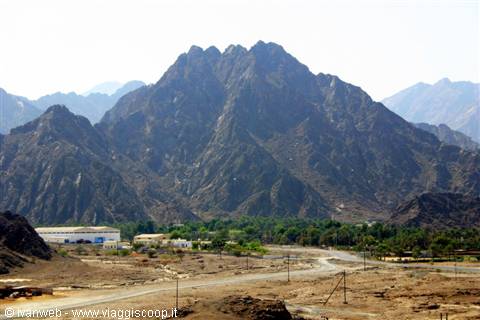Rugged rocky mountains rising above dirt road and low buildings, harsh sunlight