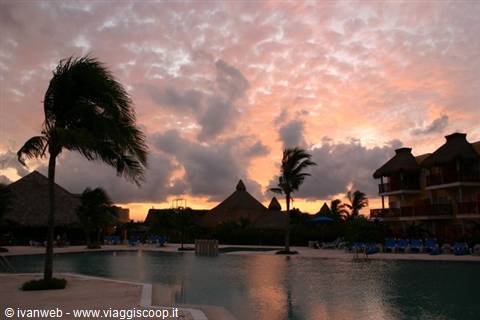 Palms and thatched roofs around the pool under pink glowing clouds