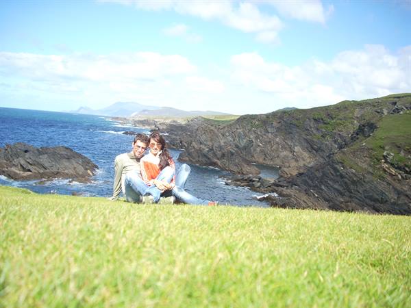 Couple sitting on grass above dark ocean cliffs, cloudy blue sky
