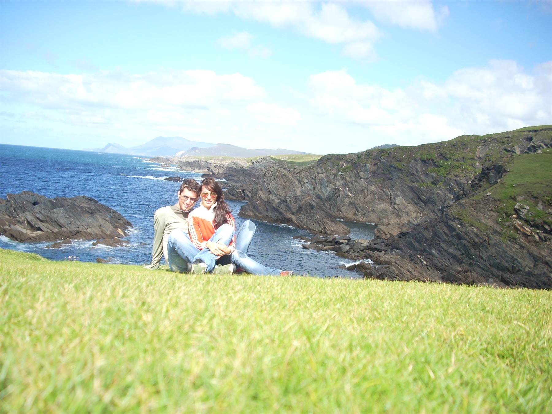 Couple sitting on grass above dark ocean cliffs, cloudy blue sky