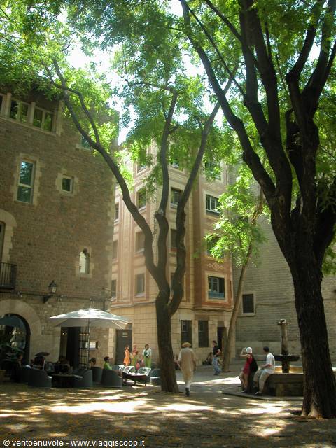 Tree-shaded square with stone fountain, café tables under umbrella and seated people