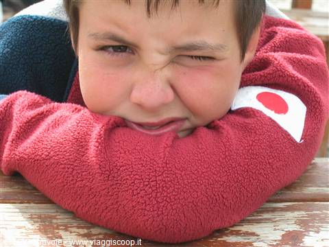 Boy in red fleece leaning on wooden table, grimacing in the sun