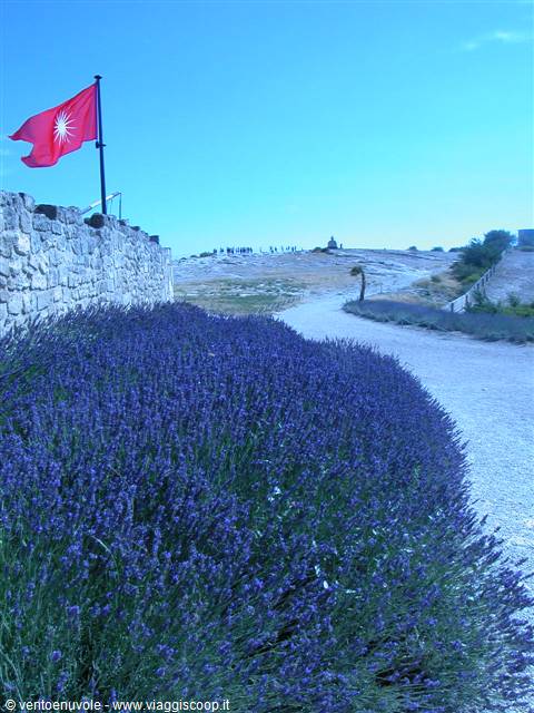 Lavender bushes below stone walls and a red flag under blue sky
