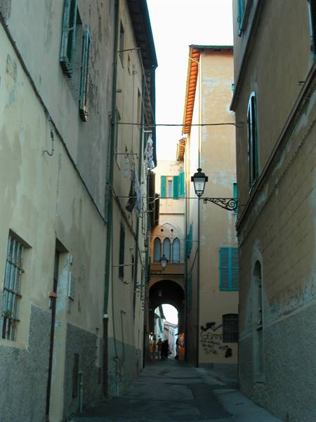 Narrow alley with ochre houses, green shutters, street lamp, people under the arch