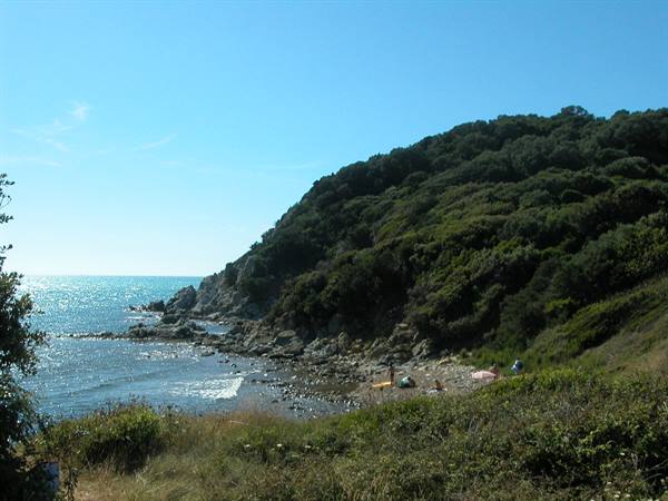 Pebbly beach with sunbathers below a green hill, blue sea and rocks