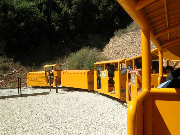 Yellow tourist train with passengers rounding gravel curve, green trees behind