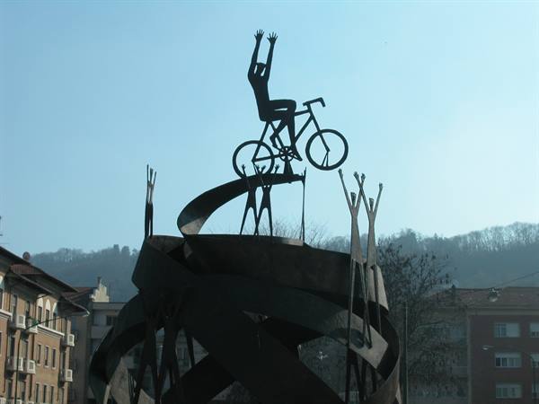 Iron sculpture of a cyclist with raised arms on a spiral, city backdrop
